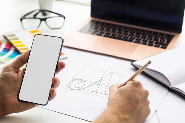 Persoon die logo tekent op papier met telefoon in de hand en laptop op achtergrond